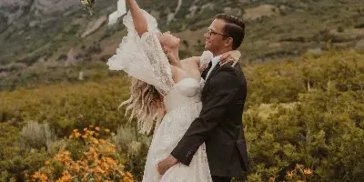 A bride and groom in a desert landscape with a mountain backdrop, the bride holding a bouquet aloft.