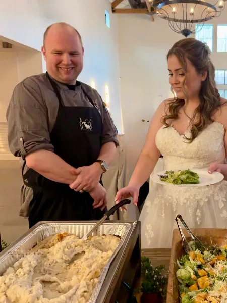 A chef in a black apron serving a plate of salad to a bride in a white wedding dress at a catering station.