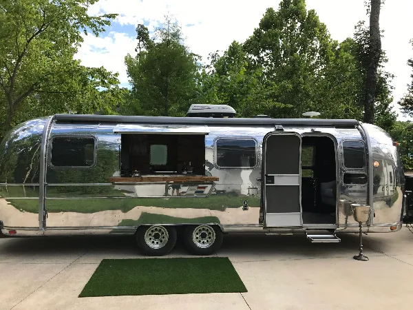 A silver Airstream trailer converted into a mobile bar and lounge with an open service window and door.