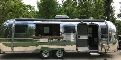 A silver Airstream trailer converted into a mobile bar and lounge with an open service window and door.