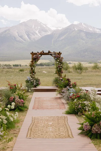A rustic wooden wedding arch decorated with wildflowers and greenery, set in a field with a mountain backdrop.