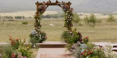 A rustic wooden wedding arch decorated with wildflowers and greenery, set in a field with a mountain backdrop.