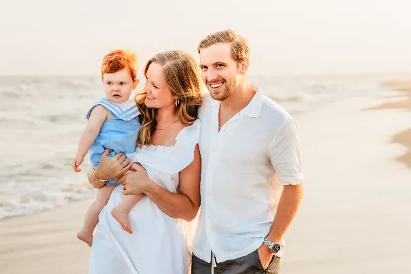 A smiling couple and a toddler in a sailor outfit posing together on a sandy beach at sunset.