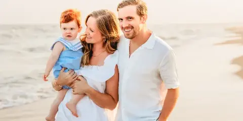 A smiling couple and a toddler in a sailor outfit posing together on a sandy beach at sunset.
