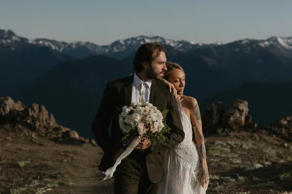 A bride and groom standing together on a rocky mountain summit with a snow-capped mountain range in the background.