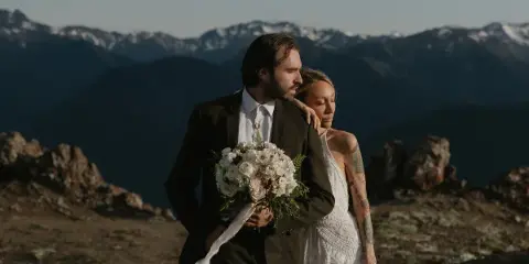 A bride and groom standing together on a rocky mountain summit with a snow-capped mountain range in the background.