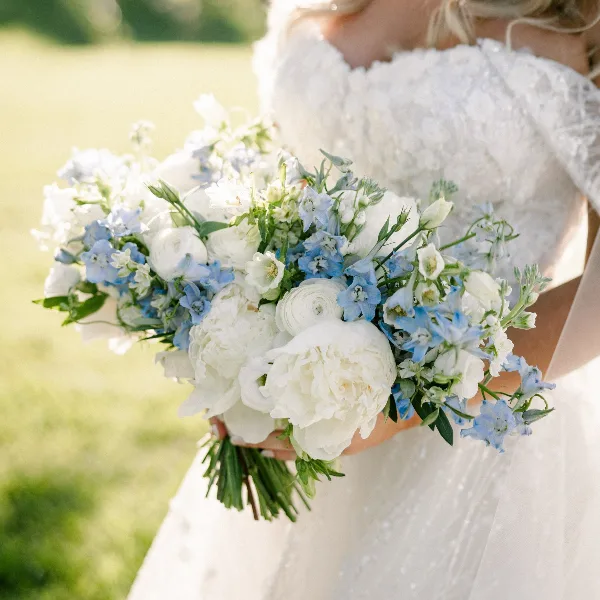 A bride in a white lace dress holding a large bouquet of white peonies and blue delphiniums.