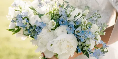 A bride in a white lace dress holding a large bouquet of white peonies and blue delphiniums.