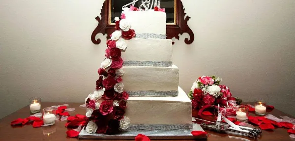A four-tier white wedding cake decorated with cascading red and white roses, displayed on a table with candles and a bouquet.