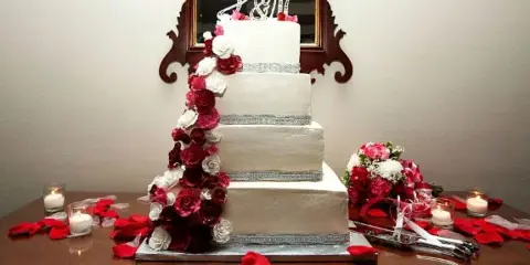 A four-tier white wedding cake decorated with cascading red and white roses, displayed on a table with candles and a bouquet.