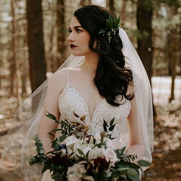 A bride in a white lace wedding dress and veil holding a bouquet in a wooded setting.