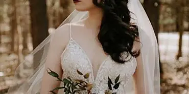 A bride in a white lace wedding dress and veil holding a bouquet in a wooded setting.