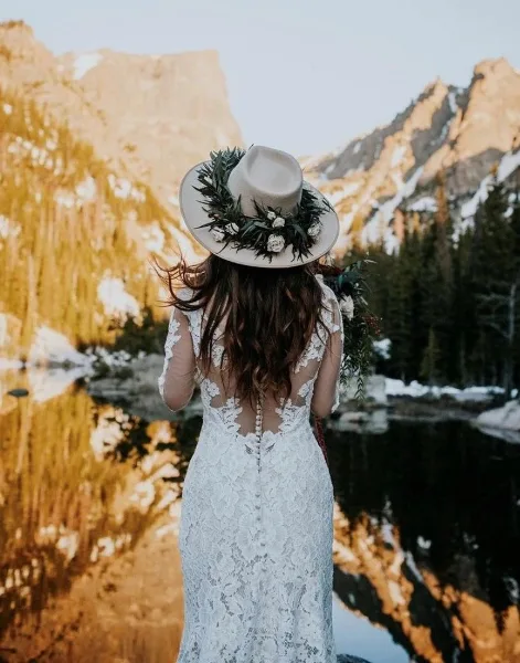 A bride in a white lace dress and floral hat standing with her back to the camera, facing a mountain lake.