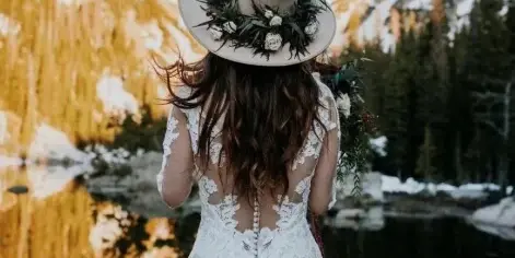 A bride in a white lace dress and floral hat standing with her back to the camera, facing a mountain lake.