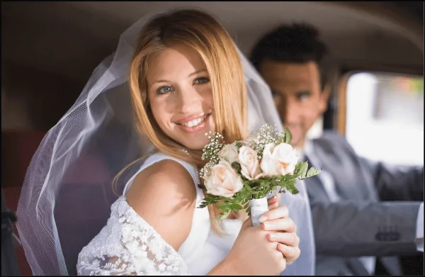 A bride holding a bouquet and a groom seated inside a car.