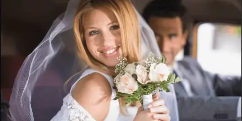 A bride holding a bouquet and a groom seated inside a car.