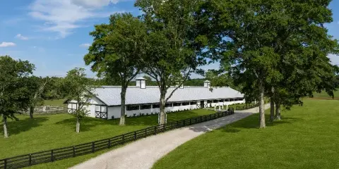 Exterior view of the Grand Pavilion at Dogwood Reserve, a white barn-style structure with a metal roof and cupolas, situated on a green lawn with a gravel driveway and black fencing.