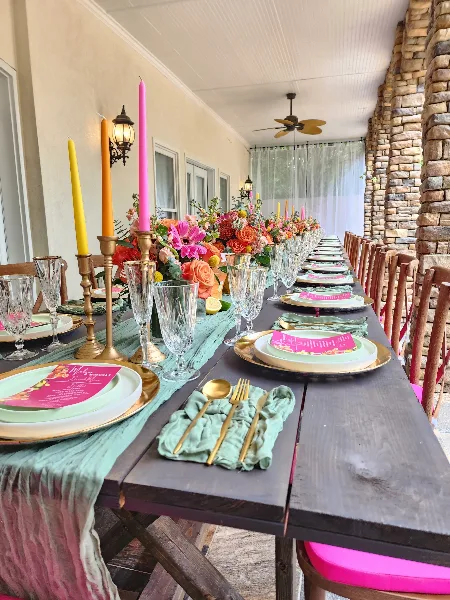 A long wooden event table set with mint green linens, gold flatware, crystal glassware, tall colorful taper candles, and a vibrant floral centerpiece.