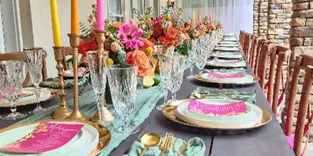 A long wooden event table set with mint green linens, gold flatware, crystal glassware, tall colorful taper candles, and a vibrant floral centerpiece.