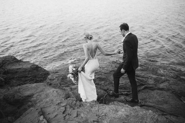 Black and white photograph of a bride and groom walking on a rocky shoreline near a large body of water.