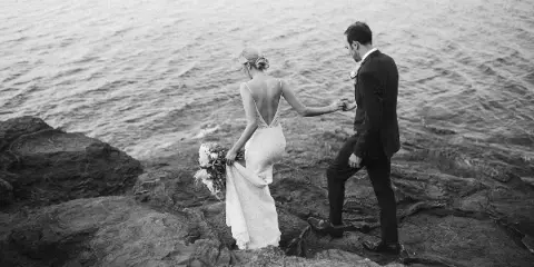 Black and white photograph of a bride and groom walking on a rocky shoreline near a large body of water.