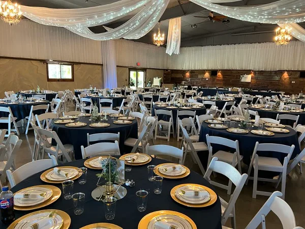 Interior of a wedding hall with round tables set with dark blue tablecloths, white chairs, and overhead drapery.