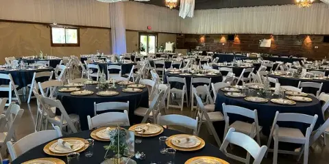 Interior of a wedding hall with round tables set with dark blue tablecloths, white chairs, and overhead drapery.