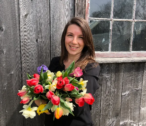 A smiling woman holding a large bouquet of colorful tulips and daffodils in front of a rustic wooden barn wall.