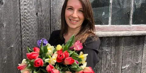 A smiling woman holding a large bouquet of colorful tulips and daffodils in front of a rustic wooden barn wall.
