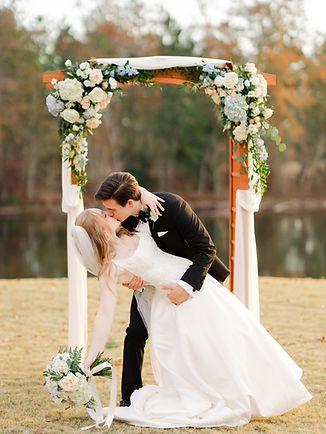A bride and groom kissing under a floral wedding arch with a bouquet in hand.