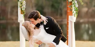 A bride and groom kissing under a floral wedding arch with a bouquet in hand.