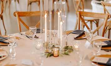 A close-up of an elegantly set wedding reception table featuring white linens, glassware, and a tall glass candle centerpiece.