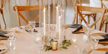 A close-up of an elegantly set wedding reception table featuring white linens, glassware, and a tall glass candle centerpiece.