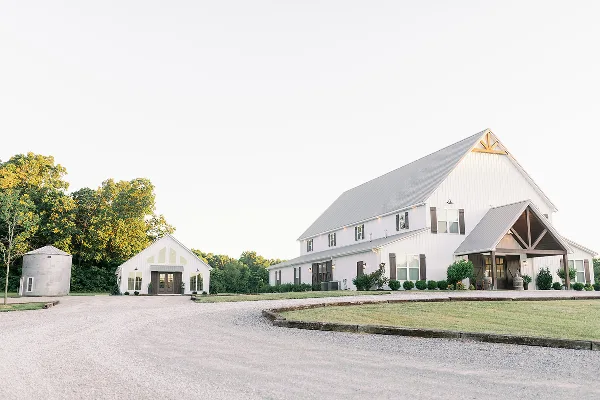 Exterior view of a large white barn wedding venue with a smaller white building and silo on a gravel driveway.