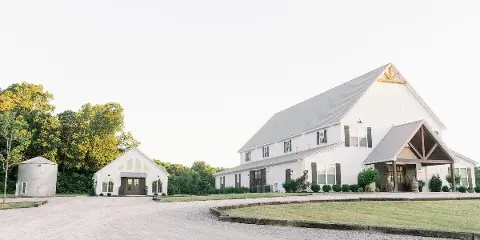 Exterior view of a large white barn wedding venue with a smaller white building and silo on a gravel driveway.