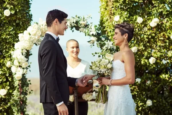 A wedding officiant in white stands behind a couple exchanging rings at a floral arch during a ceremony.