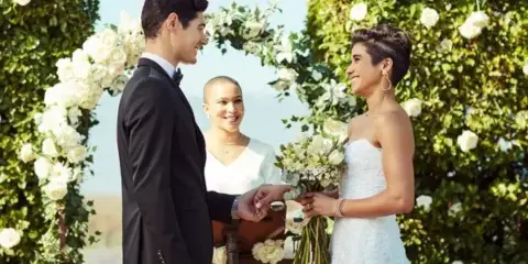 A wedding officiant in white stands behind a couple exchanging rings at a floral arch during a ceremony.
