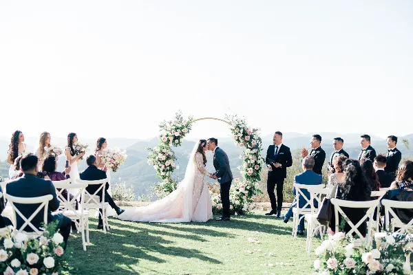 A wedding ceremony taking place outdoors on a grassy hillside with a floral arch, where a bride and groom are kissing while an officiant stands to the side.