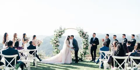 A wedding ceremony taking place outdoors on a grassy hillside with a floral arch, where a bride and groom are kissing while an officiant stands to the side.