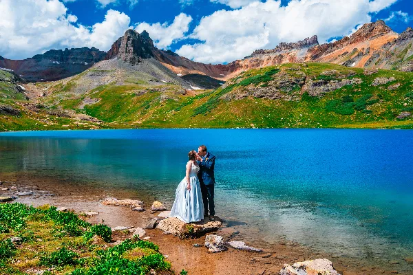 A bride and groom standing on a rocky shore at a turquoise alpine lake with rugged mountains in the background.