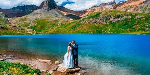 A bride and groom standing on a rocky shore at a turquoise alpine lake with rugged mountains in the background.