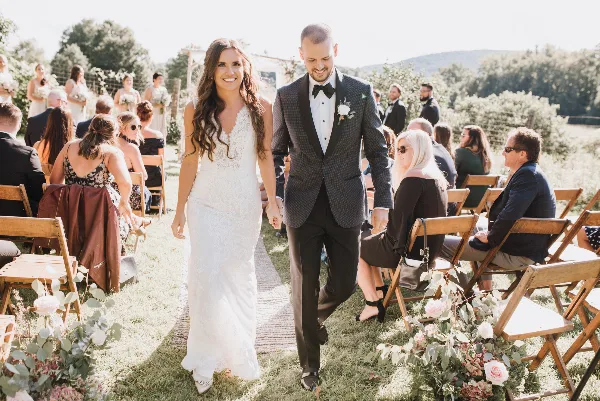 A bride and groom walking down an outdoor aisle during a wedding ceremony with seated guests.