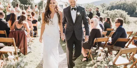 A bride and groom walking down an outdoor aisle during a wedding ceremony with seated guests.