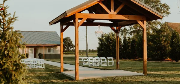 Outdoor wooden wedding ceremony pavilion with white chairs and a chandelier, set on a lawn with a barn building in the background.