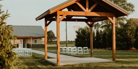 Outdoor wooden wedding ceremony pavilion with white chairs and a chandelier, set on a lawn with a barn building in the background.