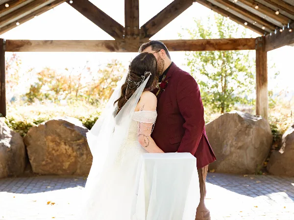 A bride and groom sharing a kiss under a wooden pavilion structure during a wedding ceremony.