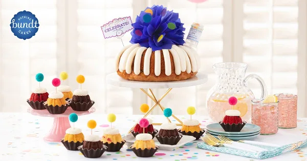 A decorated Bundt cake on a stand surrounded by assorted Bundtlets and party supplies.