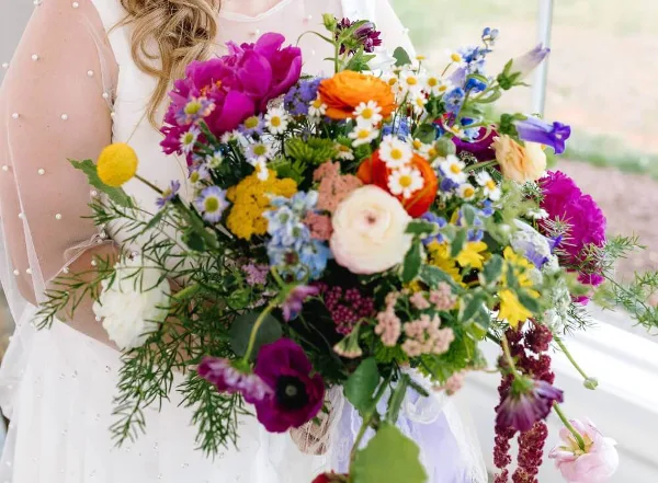 A bride holding a colorful, lush garden-style wedding bouquet featuring magenta, orange, and yellow flowers.