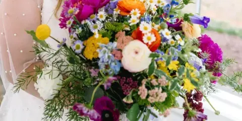 A bride holding a colorful, lush garden-style wedding bouquet featuring magenta, orange, and yellow flowers.