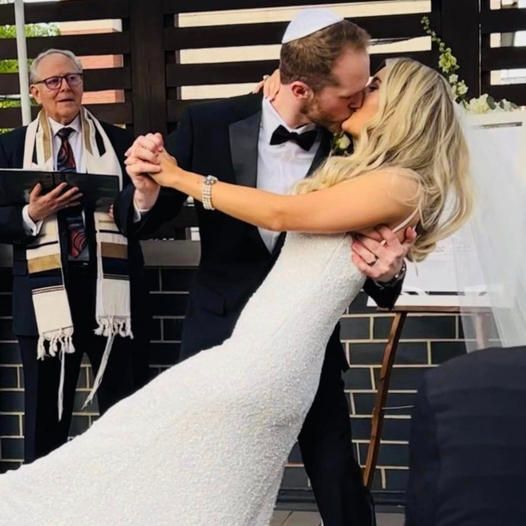 A bride and groom kiss during their wedding ceremony while an officiant in a tallit and kippah looks on.